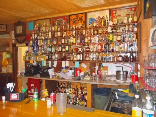 A rustic bar with a wooden counter, featuring a wall of various liquor bottles and a cluttered workspace behind it.