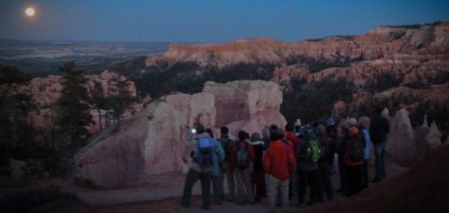 A group of people stands on a rocky overlook at dusk, watching a full moon rise over a canyon landscape.