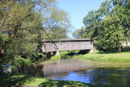 A wooden covered bridge spans a calm stream, surrounded by lush greenery and trees under a clear blue sky.