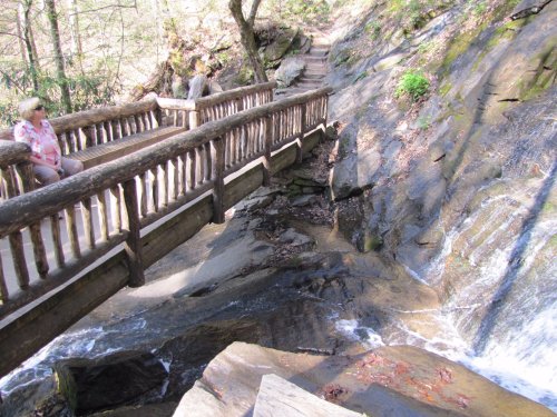 A wooden bridge over a rocky stream in a forested area, with a person sitting nearby enjoying the scenery.