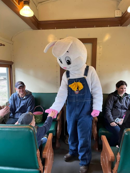 A person in a large bunny costume greets a child on a train, surrounded by smiling adults.