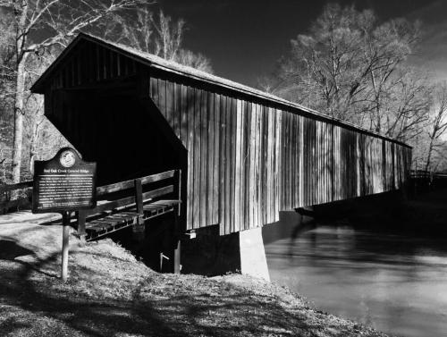 A historic covered bridge stands over a calm river, surrounded by bare trees, with a sign nearby. Black and white image.