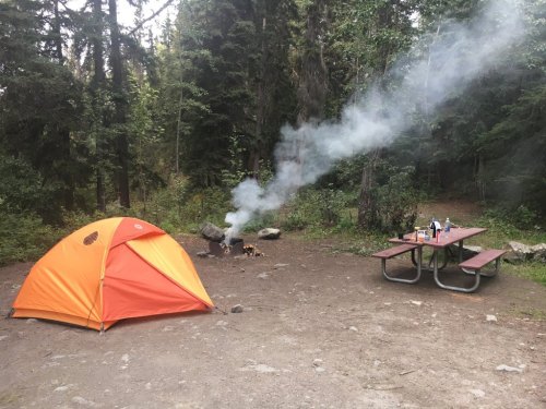 A bright orange tent near a picnic table, with smoke rising from a campfire in a wooded campsite.