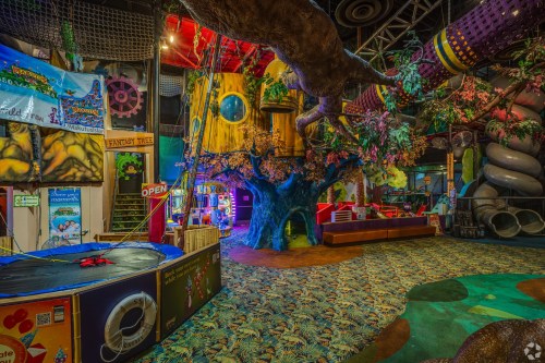 Colorful indoor play area featuring a large tree, climbing structures, and various play equipment.