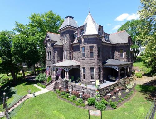 A large, historic stone mansion with a turret, surrounded by greenery and a well-maintained garden on a sunny day.
