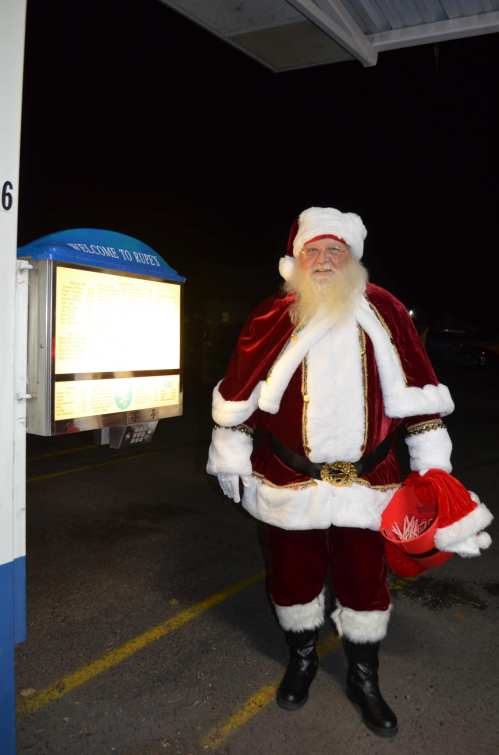 Santa Claus in a red suit and hat stands beside a menu board at night, smiling warmly.
