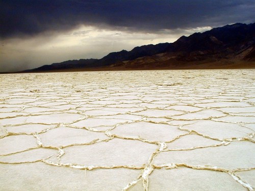 A vast, cracked salt flat under a dark, cloudy sky with distant mountains in the background.