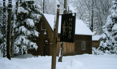 A snowy landscape featuring a wooden cabin and a sign that reads "MANKO," surrounded by trees.