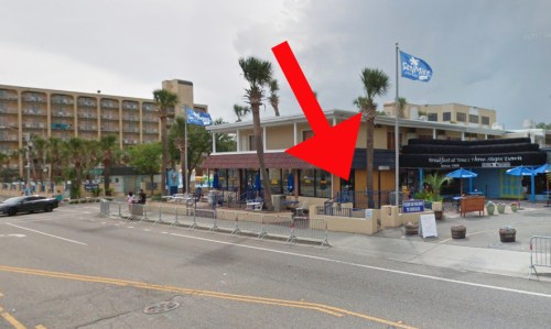 A street view of a restaurant with outdoor seating, palm trees, and a large arrow pointing to the entrance.