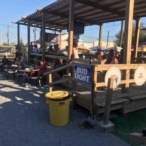 Outdoor venue with people dining, wooden seating, and a yellow trash bin. Signs for Bud Light and food options visible.