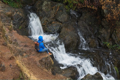 A person in a blue raincoat sits on rocks by a flowing stream, surrounded by greenery and rocky terrain.