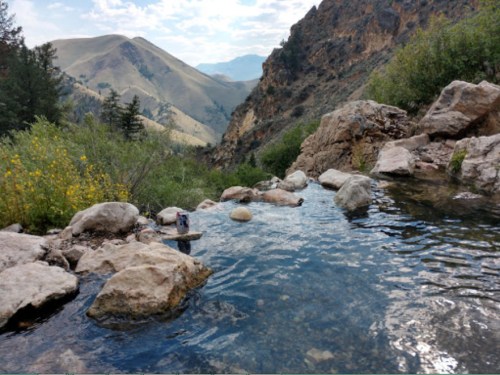 A serene natural hot spring surrounded by rocky terrain and lush greenery, with mountains in the background.