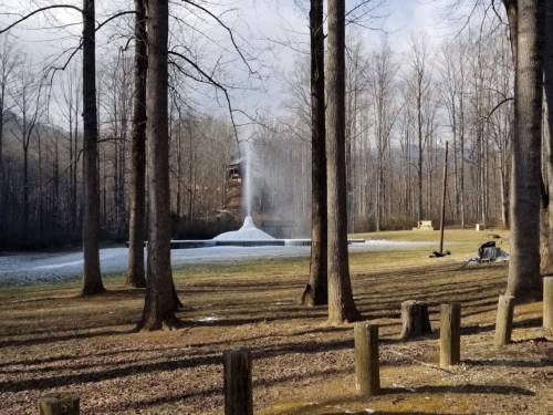 A fountain sprays water in a wooded area, surrounded by trees and a grassy field under a cloudy sky.