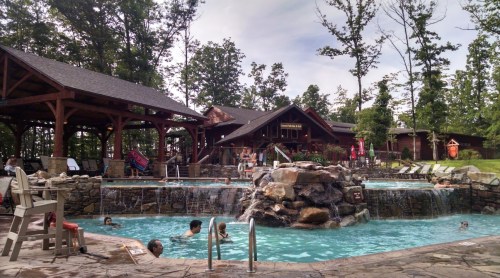 A scenic pool area with a stone waterfall, surrounded by trees and a lodge, with people enjoying the water.