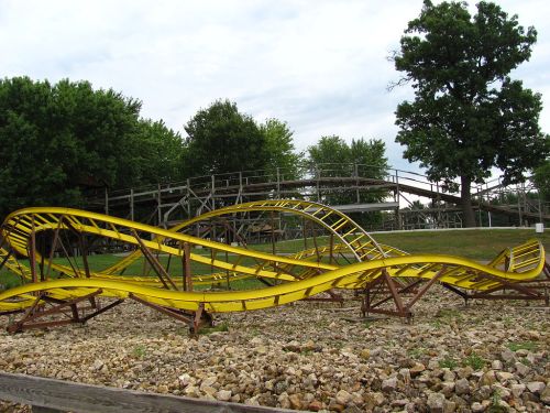 A yellow roller coaster track with twists and turns, set against a backdrop of green trees and a cloudy sky.