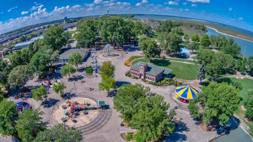 Aerial view of a park featuring playgrounds, a carousel, and green trees near a body of water under a blue sky.