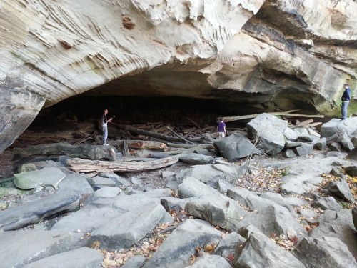 Two people explore a rocky cave entrance with wooden debris and large boulders scattered on the ground.