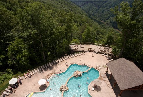 Aerial view of a pool area surrounded by trees, with people swimming and lounging on deck chairs.