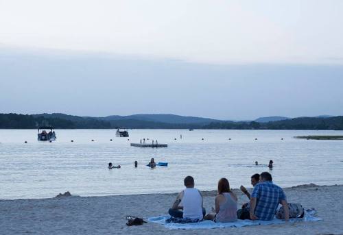 A group of people sitting on a beach at dusk, with boats on the water and others swimming nearby.