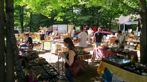 A bustling outdoor market with various handmade crafts and goods, surrounded by trees and people browsing.