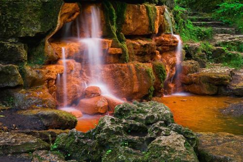 A serene waterfall cascades over moss-covered rocks into a tranquil pool, surrounded by lush greenery.