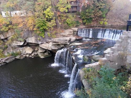 A serene waterfall cascading over rocks into a calm river, surrounded by autumn foliage and rustic buildings.