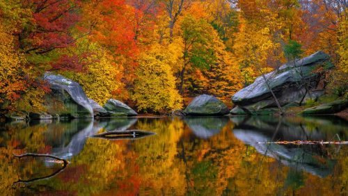 Vibrant autumn foliage reflects in a calm lake, surrounded by large rocks and trees in shades of red, orange, and yellow.