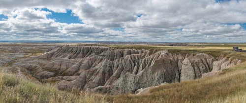 A panoramic view of rugged, colorful badlands under a cloudy sky, with grassy areas in the foreground.