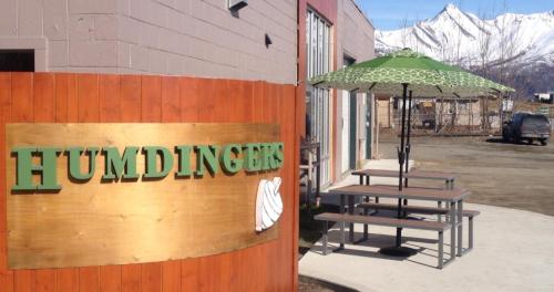 Sign for "Humdingers" with a picnic table and umbrella outside, set against a backdrop of snow-capped mountains.