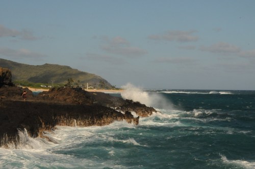 Waves crash against rocky shore with a beach and hills in the background under a clear blue sky.