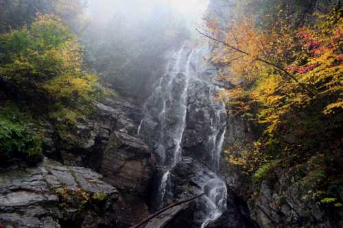 A misty waterfall cascades down rocky cliffs, surrounded by vibrant autumn foliage in a serene forest setting.