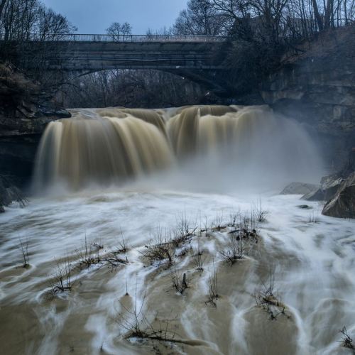 A waterfall cascades under a bridge, surrounded by trees and rocky terrain, with a misty atmosphere.