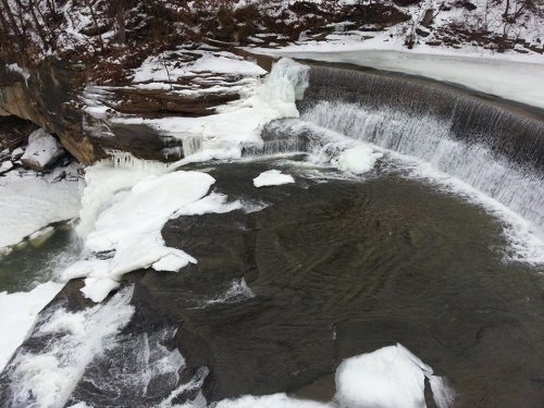 A frozen waterfall with ice formations and snow-covered rocks, surrounded by a winter landscape.