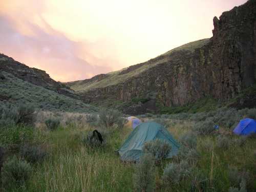 A serene camping scene with tents nestled in a grassy valley surrounded by rocky hills and a colorful sunset sky.