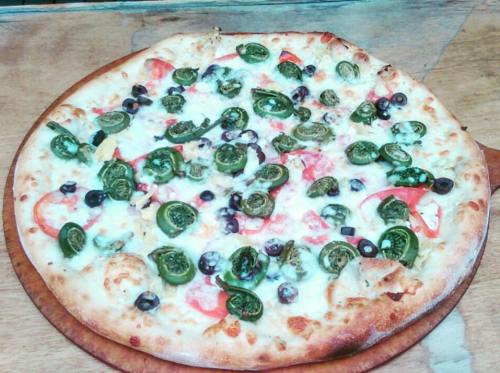 A freshly baked pizza topped with green ferns, olives, tomatoes, and melted cheese on a wooden serving board.
