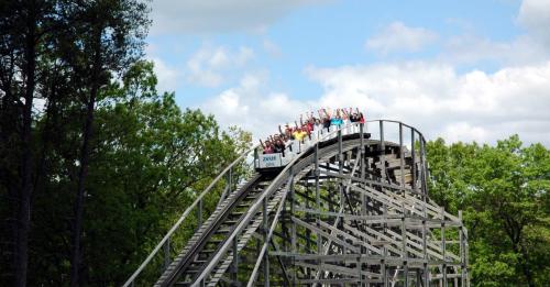 A wooden roller coaster with riders raising their hands, climbing a steep hill against a backdrop of trees and blue sky.