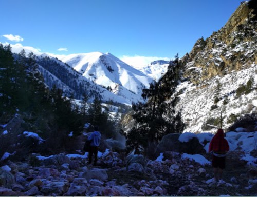 Two hikers walk along a rocky path in a snowy mountain landscape under a clear blue sky.