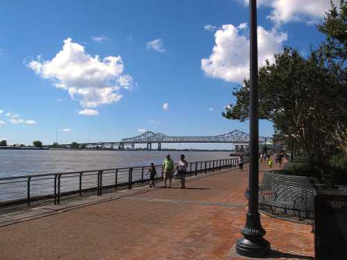 A riverside walkway with people strolling, featuring a bridge in the background under a blue sky with fluffy clouds.