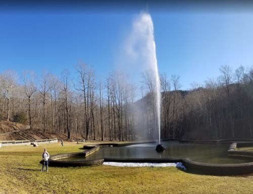 A serene landscape featuring a fountain in a pond, surrounded by bare trees and a clear blue sky.