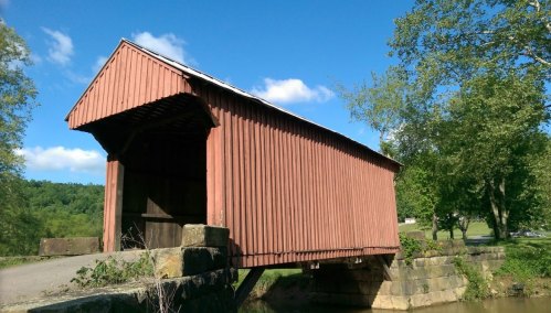 A red covered bridge spans a small stream, surrounded by green trees and a clear blue sky.