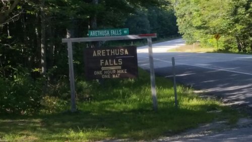 Sign for Arethusa Falls with directions for a one-hour hike, surrounded by trees and a winding road.