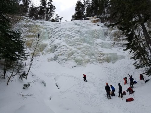 A snowy landscape featuring a frozen waterfall, with climbers and people preparing gear at the base.