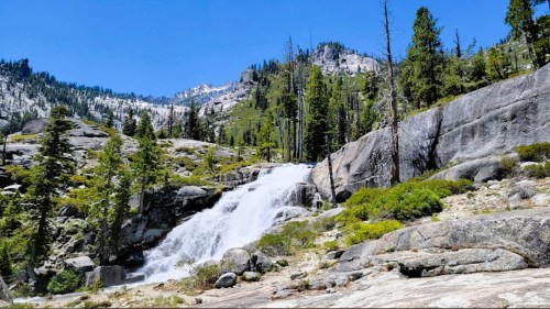 A scenic waterfall cascades over rocks, surrounded by lush greenery and towering mountains under a clear blue sky.