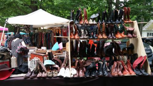 A display of various styles of boots and shoes at an outdoor market, with colorful clothing in the background.