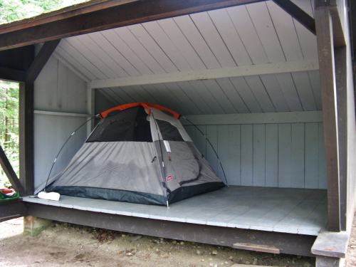 A gray tent with an orange top set up inside a wooden shelter with a slanted roof.