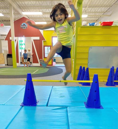 A young girl jumps over blue cones in a colorful indoor play area while two children watch in the background.