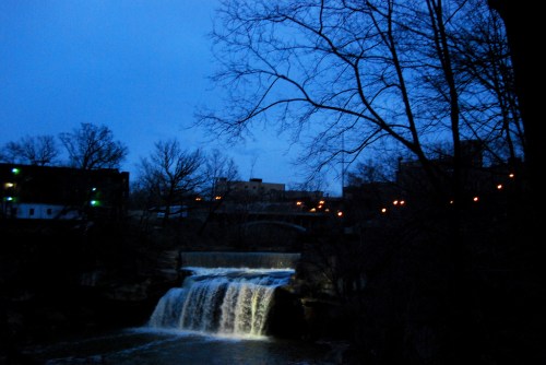 A waterfall cascades under a twilight sky, with silhouetted trees and distant city lights in the background.