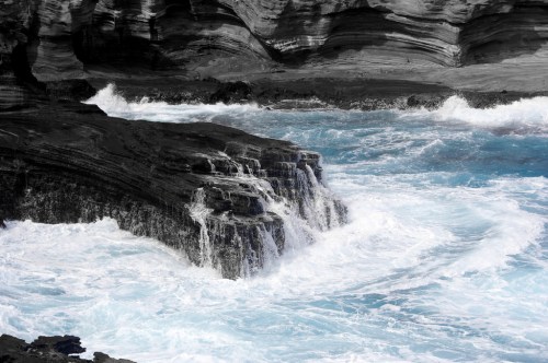 Waves crash against rocky cliffs, creating white foam against a backdrop of dark, layered rock formations.