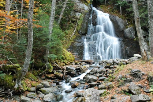A serene waterfall cascades over rocks, surrounded by lush greenery and autumn foliage.