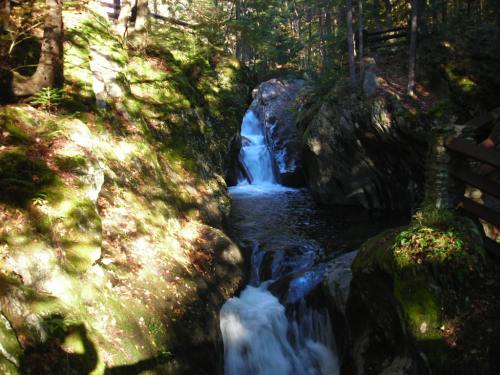 A serene waterfall cascades through rocky terrain, surrounded by lush greenery and dappled sunlight.